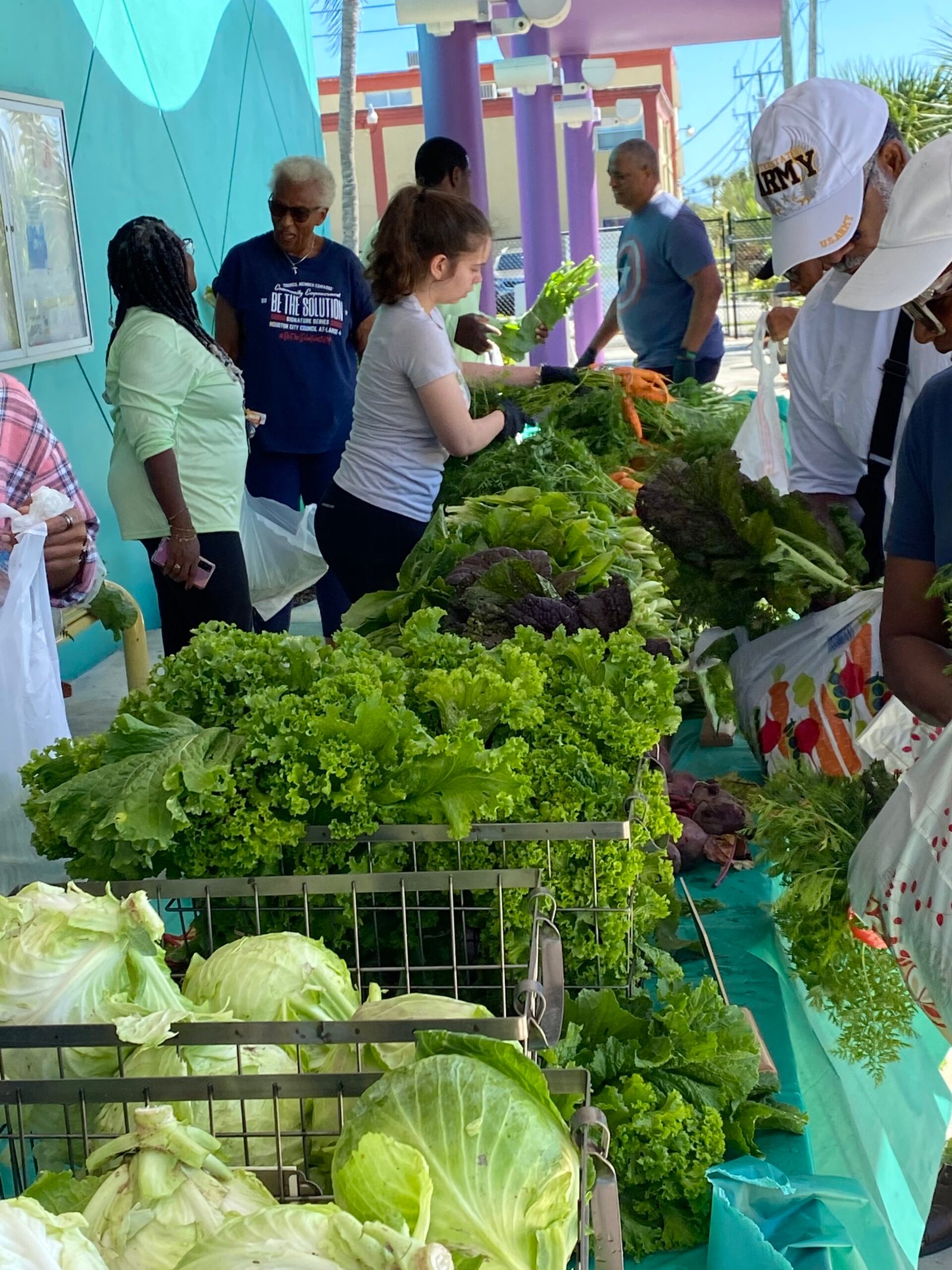 Donate food near me - Volunteers and community members gather at a fresh produce distribution event, with tables full of leafy greens, cabbages, and carrots.