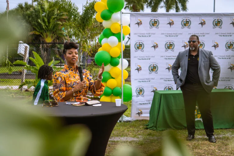 A woman speaks into a microphone at an outdoor event, seated at a black table with balloons in green, yellow, and white behind her, while a man in a suit stands nearby in front of a JFK Middle School event banner.