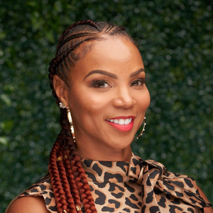 Headshot of Veleké Brown, smiling, with braided hair and wearing a leopard print top against a green leafy background.