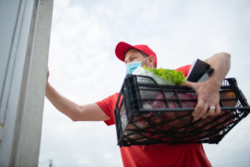 A man in a red shirt and cap delivers groceries with safety measures. Outdoors.